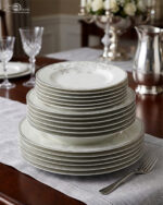 A stacked 18-piece elegant white super bone china dinner plate set featuring a delicate grey floral pattern on the rims, neatly arranged on a grey fabric runner over a wooden dining table with crystal glasses and a silver pitcher in the background.