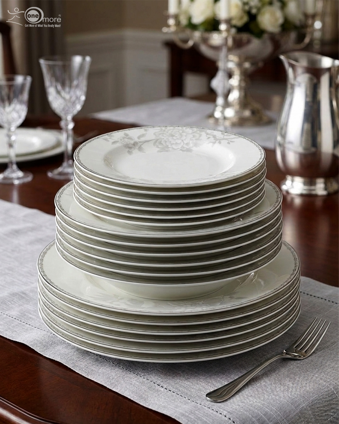 5 A stacked 18-piece elegant white super bone china dinner plate set featuring a delicate grey floral pattern on the rims, neatly arranged on a grey fabric runner over a wooden dining table with crystal glasses and a silver pitcher in the background.