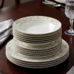 A stacked 18-piece premium super bone china dinnerware set with elegant floral and striped rim borders, neatly arranged on a dark wooden dining table next to a crystal glass and fabric napkin