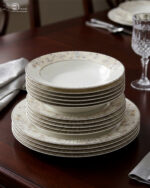 A stacked 18-piece premium super bone china dinnerware set with elegant floral and striped rim borders, neatly arranged on a dark wooden dining table next to a crystal glass and fabric napkin