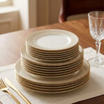 A neatly stacked 18-piece super bone china dinner plate set with elegant gold borders, arranged on a beige placemat next to a gold fork, a gold knife, and a crystal wine glass on a wooden table.