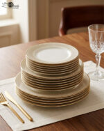 A neatly stacked 18-piece super bone china dinner plate set with elegant gold borders, arranged on a beige placemat next to a gold fork, a gold knife, and a crystal wine glass on a wooden table.