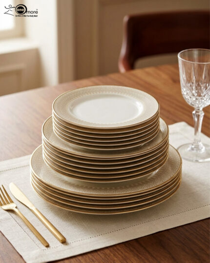A neatly stacked 18-piece super bone china dinner plate set with elegant gold borders, arranged on a beige placemat next to a gold fork, a gold knife, and a crystal wine glass on a wooden table.