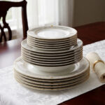 A stacked 18-piece super bone china dinner plate set featuring a white porcelain body with an elegant gold geometric border pattern, neatly arranged on a white damask table runner over a dark wooden dining table next to a rolled napkin with a gold ring.