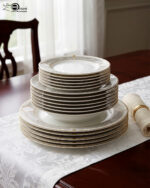 A stacked 18-piece super bone china dinner plate set featuring a white porcelain body with an elegant gold geometric border pattern, neatly arranged on a white damask table runner over a dark wooden dining table next to a rolled napkin with a gold ring.