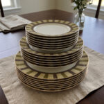 A stacked 18-piece super bone china plate set neatly arranged on a beige linen placemat over a wooden dining table. The ceramic dinnerware set features 6 quarter plates stacked on top, 6 deep soup plates in the middle, and 6 large rice plates at the bottom. Each plate showcases an elegant radiating striped pattern rim in olive green, cream, and taupe tones with fine gold rim detailing. A blur background shows a window and a small flower vase