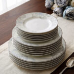 A neatly stacked 18-piece super bone china dinner plate set featuring a delicate grey and blue floral design on a beige table runner alongside a silver spoon and blue flowers.
