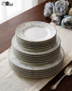 A neatly stacked 18-piece super bone china dinner plate set featuring a delicate grey and blue floral design on a beige table runner alongside a silver spoon and blue flowers.