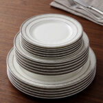 A stacked 18-piece white super bone china plate set with elegant silver-grey floral borders, displayed on a dark wooden table next to a folded cloth napkin with a knife and fork