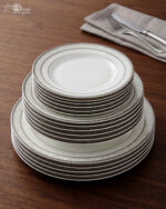 A stacked 18-piece white super bone china plate set with elegant silver-grey floral borders, displayed on a dark wooden table next to a folded cloth napkin with a knife and fork