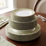 A neatly stacked 18-piece super bone china dinnerware set on a dark wooden dining table. The set includes large rice plates on the bottom, deep plates in the middle, and smaller quarter plates on top, all featuring an elegant light green and gold brushstroke pattern around the borders. Green linen napkins and silver cutlery are arranged neatly beside the stack.