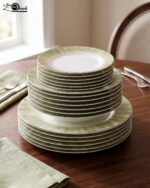 A neatly stacked 18-piece super bone china dinnerware set on a dark wooden dining table. The set includes large rice plates on the bottom, deep plates in the middle, and smaller quarter plates on top, all featuring an elegant light green and gold brushstroke pattern around the borders. Green linen napkins and silver cutlery are arranged neatly beside the stack.