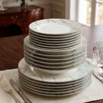 A neatly stacked 18-piece super bone china dinner plate set featuring a white and mint green marble pattern with gold veining, arranged on a dining table with silverware and a crystal glass.