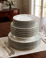 A neatly stacked 18-piece super bone china dinner plate set featuring a white and mint green marble pattern with gold veining, arranged on a dining table with silverware and a crystal glass.