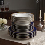 A stacked 18-piece premium super bone china dinnerware set featuring royal blue, gold rims, and geometric patterned borders, elegantly arranged on a wooden dining table with candles and roses.