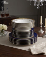 A stacked 18-piece premium super bone china dinnerware set featuring royal blue, gold rims, and geometric patterned borders, elegantly arranged on a wooden dining table with candles and roses.