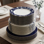 A neatly stacked 18-piece luxury dinnerware set made of Super Bone China on a wooden dining table. The stack shows three sizes of plates: the top quarter plates feature an intricate white leaf and floral pattern on a royal blue border, the middle deep plates show a subtle blue pattern, and the bottom large rice plates are a solid deep royal blue with thin gold rims. The setting is completed with elegant silverware and a small glass vase with green leaves in the background
