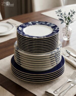 A neatly stacked 18-piece luxury dinnerware set made of Super Bone China on a wooden dining table. The stack shows three sizes of plates: the top quarter plates feature an intricate white leaf and floral pattern on a royal blue border, the middle deep plates show a subtle blue pattern, and the bottom large rice plates are a solid deep royal blue with thin gold rims. The setting is completed with elegant silverware and a small glass vase with green leaves in the background