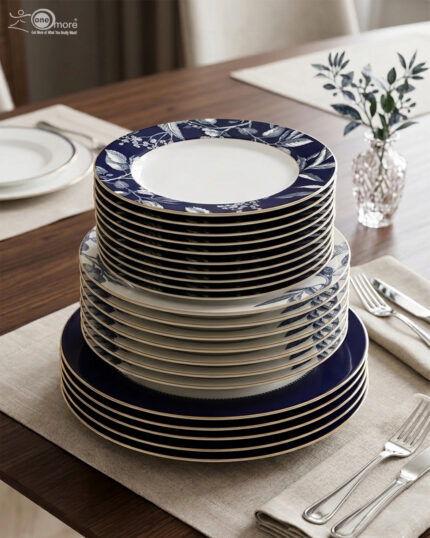 A neatly stacked 18-piece luxury dinnerware set made of Super Bone China on a wooden dining table. The stack shows three sizes of plates: the top quarter plates feature an intricate white leaf and floral pattern on a royal blue border, the middle deep plates show a subtle blue pattern, and the bottom large rice plates are a solid deep royal blue with thin gold rims. The setting is completed with elegant silverware and a small glass vase with green leaves in the background
