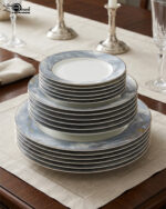 A neatly stacked 18-piece luxury super bone china plate set on a dining table runner. The dinnerware features a contemporary blue and gray ocean marble pattern with subtle gold vein accents and a thin silver rim. The stack consists of three sizes: small quarter plates on top, medium deep soup plates in the middle, and large flat rice plates at the bottom, displayed in a brightly lit, elegant dining room setting with candlesticks in the background.