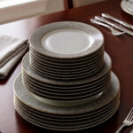 A stacked 18-piece white Super Bone China dinner plate set with elegant gold borders, arranged neatly on a polished wooden dining table alongside a wine glass and silverware under warm chandelier lighting.