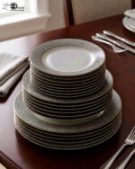 A stacked 18-piece white Super Bone China dinner plate set with elegant gold borders, arranged neatly on a polished wooden dining table alongside a wine glass and silverware under warm chandelier lighting.