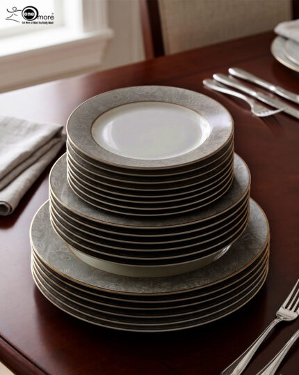 A stacked 18-piece white Super Bone China dinner plate set with elegant gold borders, arranged neatly on a polished wooden dining table alongside a wine glass and silverware under warm chandelier lighting.