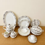 A complete Super Bone China dinner set from 6.jpg, featuring a white base with intricate grey floral borders. The set includes plates, bowls, a teapot, a tureen, and teacups displayed on a wooden surface