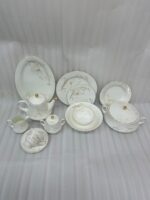 A complete super bone china dinnerware set in 7.jpg, featuring white plates, bowls, a teapot, and tea cups adorned with delicate cream floral patterns and luxurious gold-rimmed edges on a wooden surface.