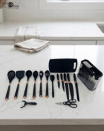 A comprehensive 17-piece kitchen tool set featuring black and rose gold silicone utensils, a matching knife set with marble-patterned handles, a vegetable peeler, kitchen shears, and a black cutting board, all stored in a clear geometric textured organizer.
