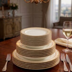 A stacked 18-piece white Super Bone China dinner plate set with elegant gold borders, arranged neatly on a polished wooden dining table alongside a wine glass and silverware under warm chandelier lighting.