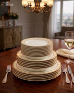A stacked 18-piece white Super Bone China dinner plate set with elegant gold borders, arranged neatly on a polished wooden dining table alongside a wine glass and silverware under warm chandelier lighting.