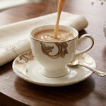 A steaming stream of milk tea being poured into a luxury white Super Bone China cup with gold-leaf designs, resting on a matching saucer with a silver spoon on a dark wooden table