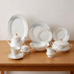 A comprehensive white super bone china dinner set from 7.jpg featuring an elegant blue leaf design and gold-rimmed edges, arranged on a wooden table. Includes plates, bowls, a teapot, cups, and a tureen.