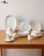 A comprehensive white super bone china dinner set from 7.jpg featuring an elegant blue leaf design and gold-rimmed edges, arranged on a wooden table. Includes plates, bowls, a teapot, cups, and a tureen.