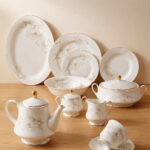 A complete super bone china dinnerware set in 7.jpg, featuring white plates, bowls, a teapot, and tea cups adorned with delicate cream floral patterns and luxurious gold-rimmed edges on a wooden surface.