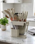 A comprehensive 16-piece kitchen tool set featuring cream-colored silicone utensils with marble-patterned handles, rose gold accents, a matching knife set, kitchen shears, a peeler, and a cutting board, all stored in a clear geometric organizer.