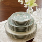 A stack of 18 super bone china plates featuring a sage green and white floral design, including rice plates, deep plates, and quarter plates, displayed on a white damask runner