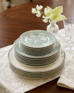 A stack of 18 super bone china plates featuring a sage green and white floral design, including rice plates, deep plates, and quarter plates, displayed on a white damask runner
