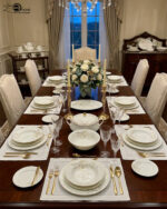 A complete super bone china dinnerware set in 7.jpg, featuring white plates, bowls, a teapot, and tea cups adorned with delicate cream floral patterns and luxurious gold-rimmed edges on a wooden surface.