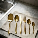 A 36-piece luxury gold cutlery set arranged on a linen cloth next to a kitchen sink, featuring intricate floral engravings on the handles, including assorted dinner spoons, tea spoons, forks, knives, and serving utensils.