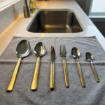 Six pieces from a 36-piece premium cutlery set featuring polished stainless steel heads and intricate, decorative gold-tone handles, displayed on a grey fabric surface near a modern kitchen sink.