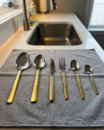 Six pieces from a 36-piece premium cutlery set featuring polished stainless steel heads and intricate, decorative gold-tone handles, displayed on a grey fabric surface near a modern kitchen sink.