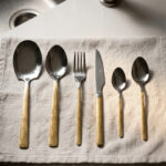Flat lay of a 36-piece stainless steel cutlery set with polished silver heads and intricately engraved gold floral-patterned handles, arranged neatly on a textured beige linen cloth.
