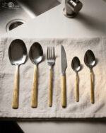 Flat lay of a 36-piece stainless steel cutlery set with polished silver heads and intricately engraved gold floral-patterned handles, arranged neatly on a textured beige linen cloth.