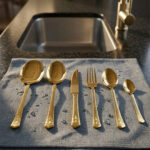 A collection of 36 gold-finished hammered cutlery pieces, including forks, knives, and various spoons, arranged neatly on a grey linen cloth beside a modern kitchen sink.