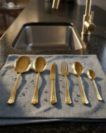 A collection of 36 gold-finished hammered cutlery pieces, including forks, knives, and various spoons, arranged neatly on a grey linen cloth beside a modern kitchen sink.