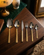 High-end 36-piece stainless steel cutlery set featuring polished silver heads and gold handles with a geometric Greek key pattern, arranged elegantly on a dark wooden table.