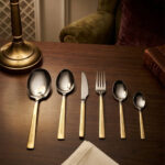 A 36-piece luxury One More cutlery set featuring elegant gold-accented handles with a geometric Greek key pattern, arranged professionally on a dark wood dining table.