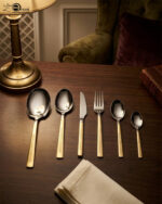 A 36-piece luxury One More cutlery set featuring elegant gold-accented handles with a geometric Greek key pattern, arranged professionally on a dark wood dining table.